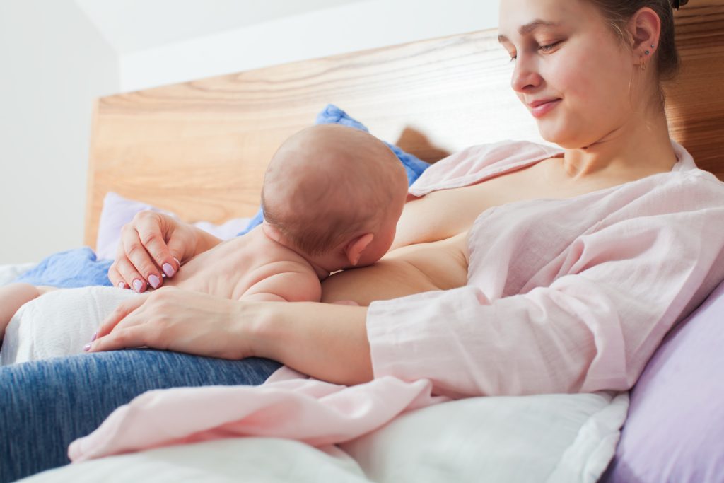 young mother half laying bed supported by pillows holding newborn baby skin skin her stomach learning breastfeed with baby led attachment biological nurturing